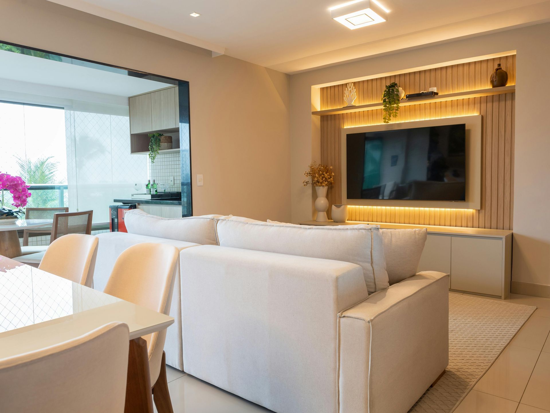 A bright modern living room with a cream sofa, a mounted television in a wood-paneled feature wall, and a dining area.