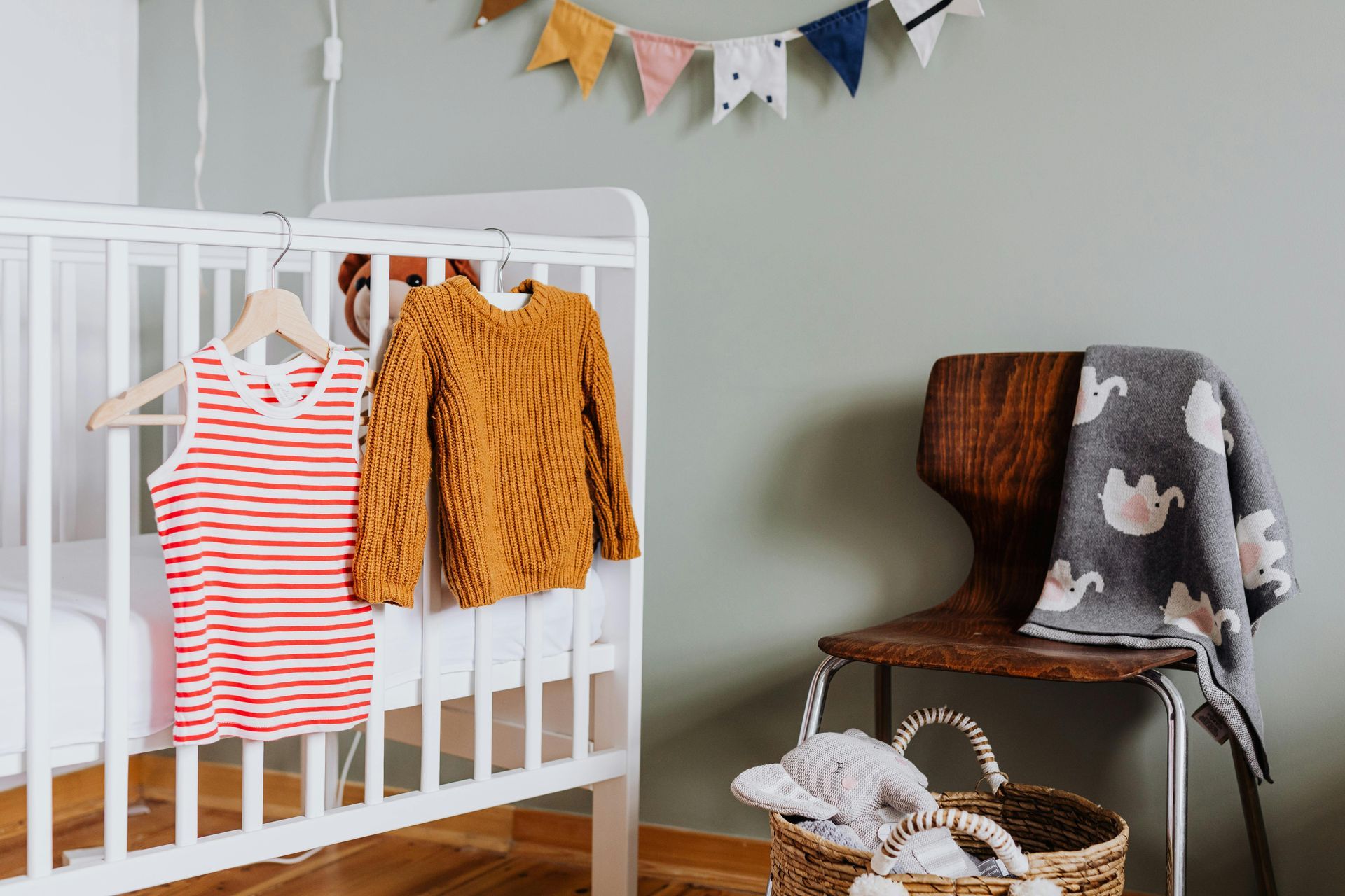A nursery with a white crib, clothes hanging on it, a blanket on a chair, and a basket with toys.