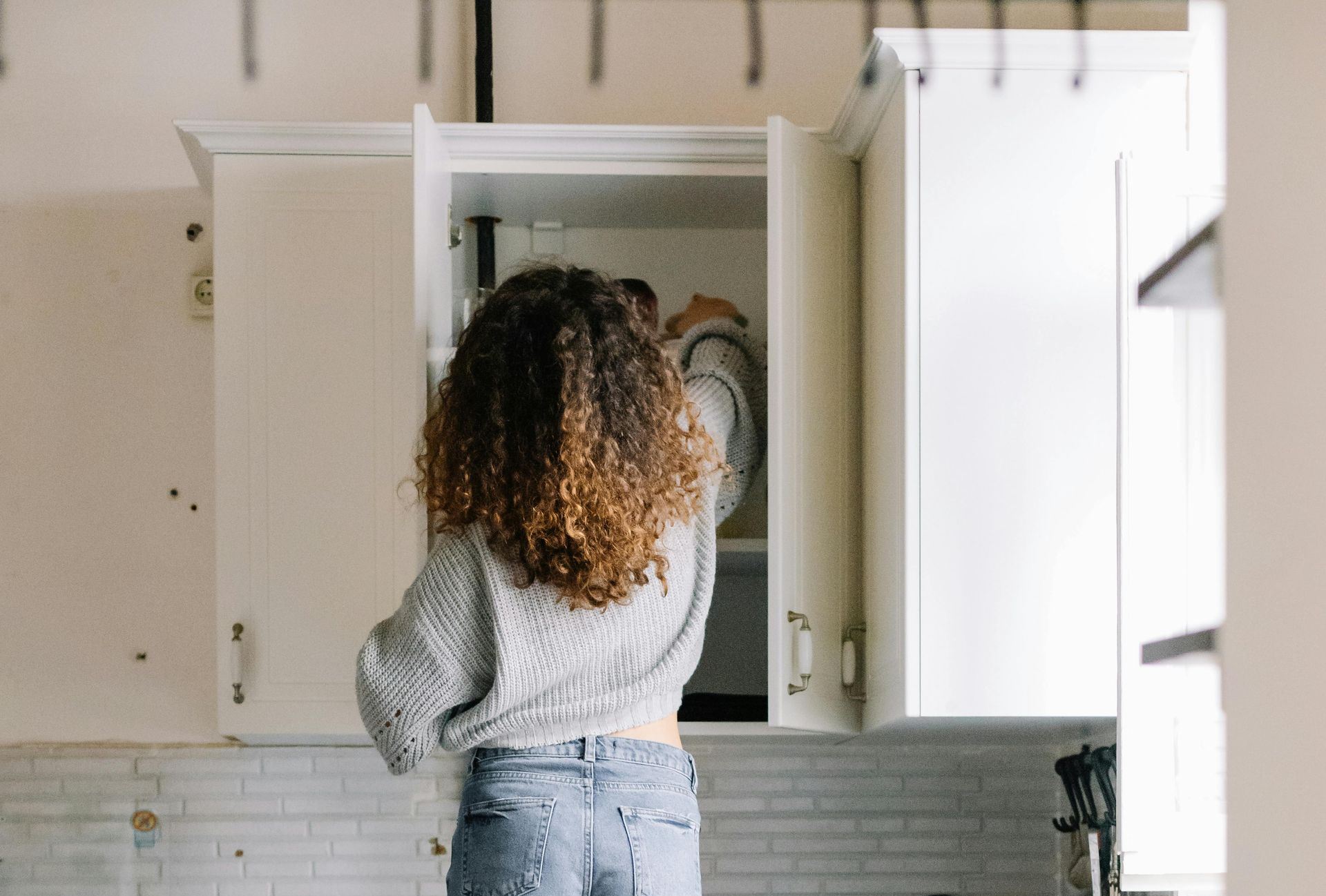 A person with curly hair wearing a grey sweater reaches into an open white kitchen cabinet to retrieve a white plate.