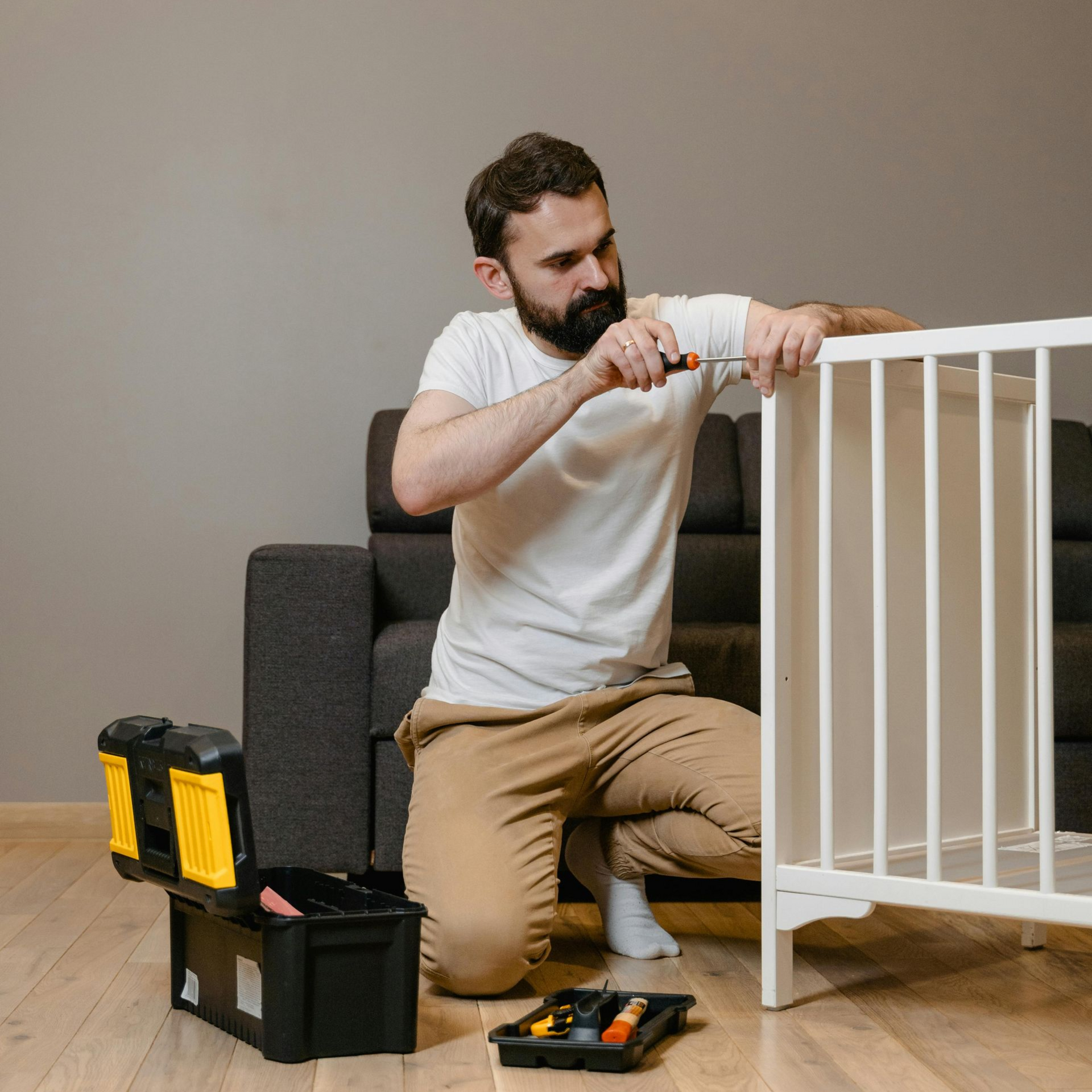 Man kneeling, assembling a white crib with tools in a toolbox on a hardwood floor.