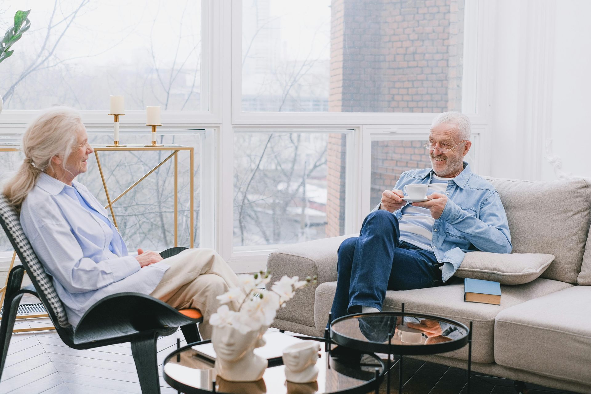 Two people sit in a bright, modern living room, conversing while one holds a teacup.