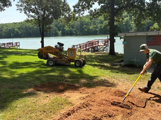 Person raking dirt by a lakeside, with a yellow utility cart and dock in the background.