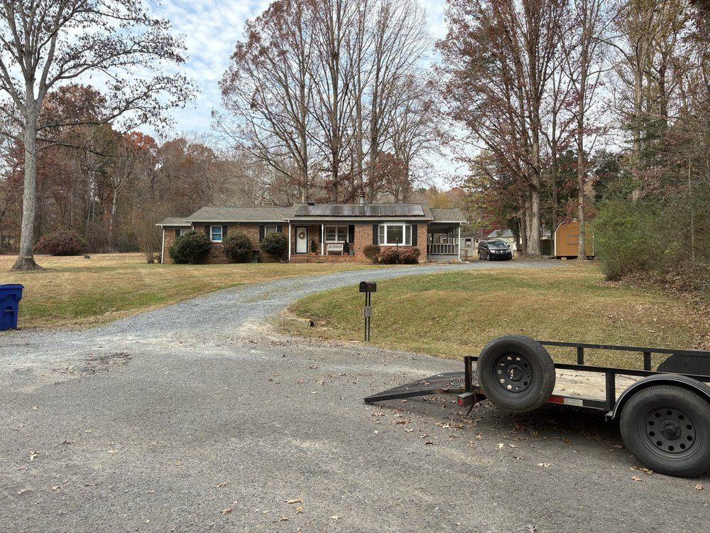 Ranch house with a gravel driveway, trailer parked in the foreground, trees in the background under an overcast sky.