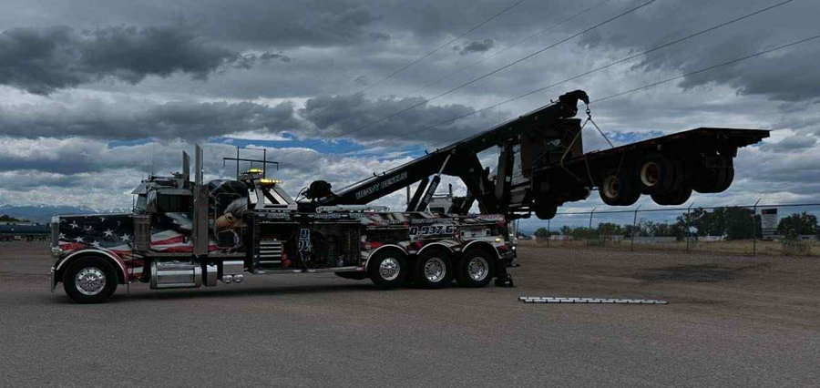 A tow truck lifting a flatbed trailer against a cloudy sky.