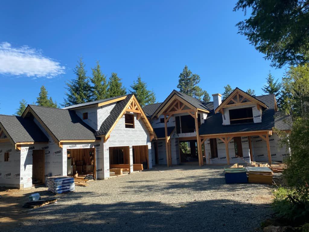 House under construction with exposed timber framing and dark shingle roof, on a gravel lot under a blue sky.