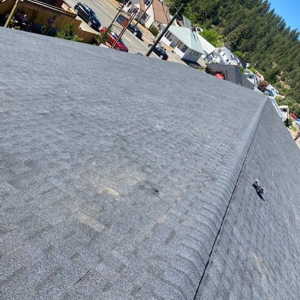 View from above of a dark grey asphalt shingle roof. Buildings, road, and trees are in the background.