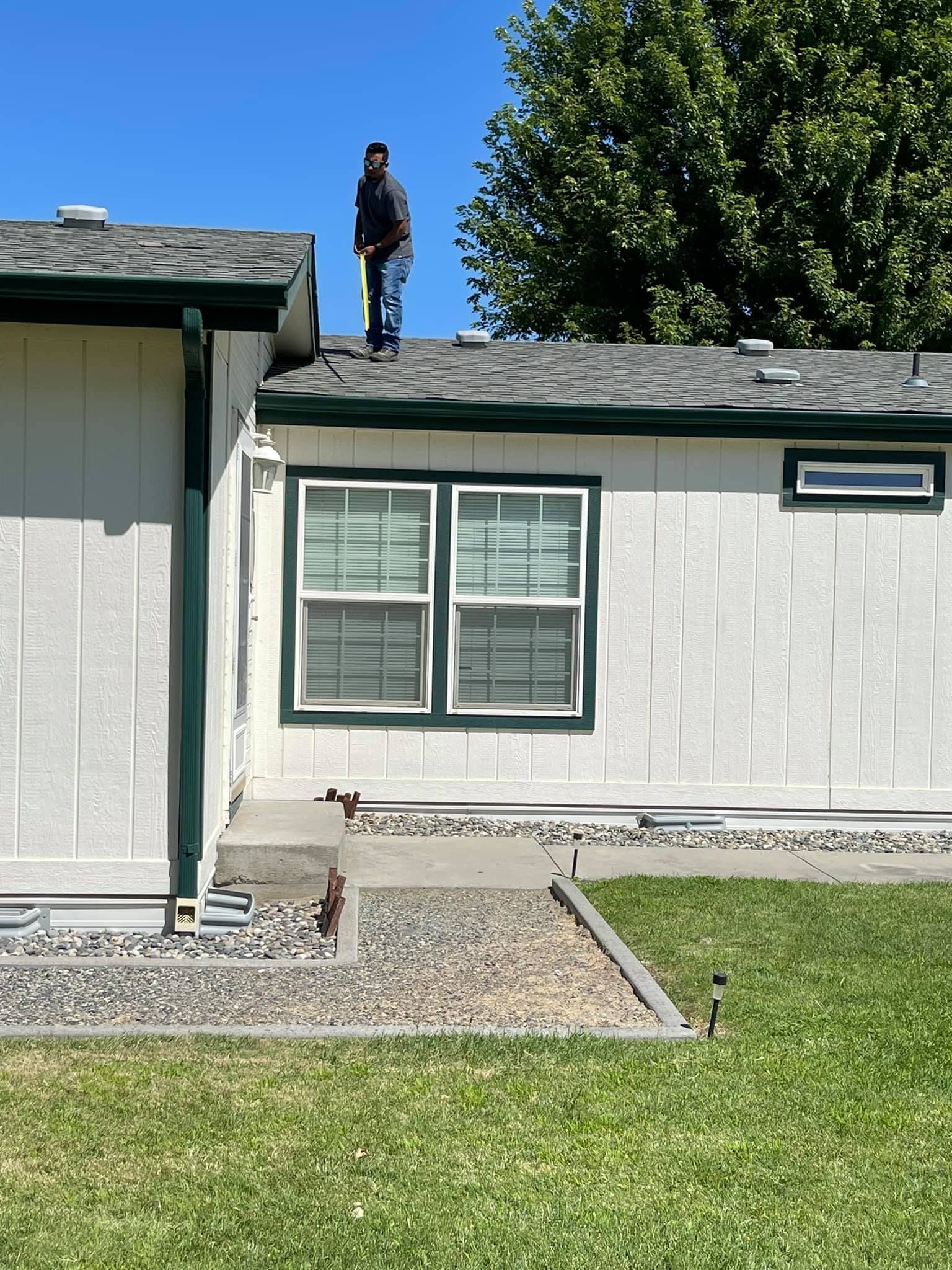 Person standing on a house roof near a chimney. Green trim and grass with blue sky.