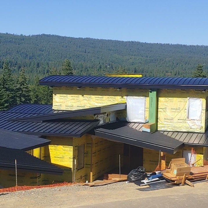 House under construction with black roof, yellow exterior wrap, and a forest background.