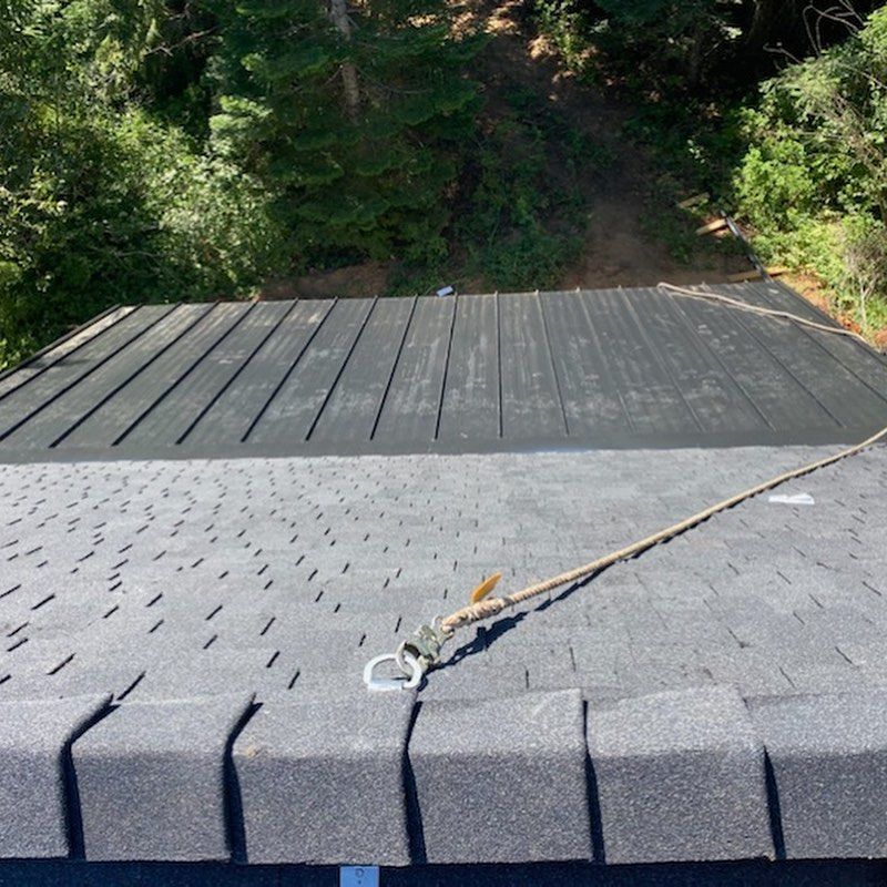 View of a dark shingle roof with a safety rope secured to it, with trees in the background.