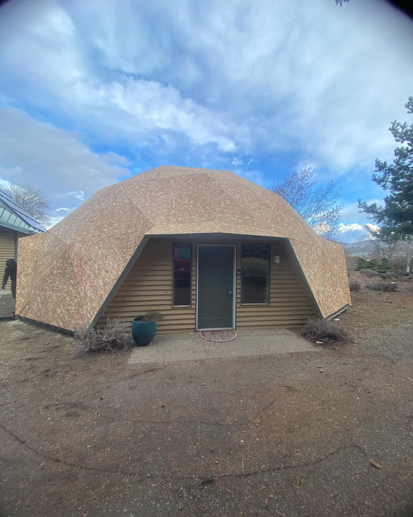 Geodesic dome house with wooden siding and a dark green door, on a dirt lot.