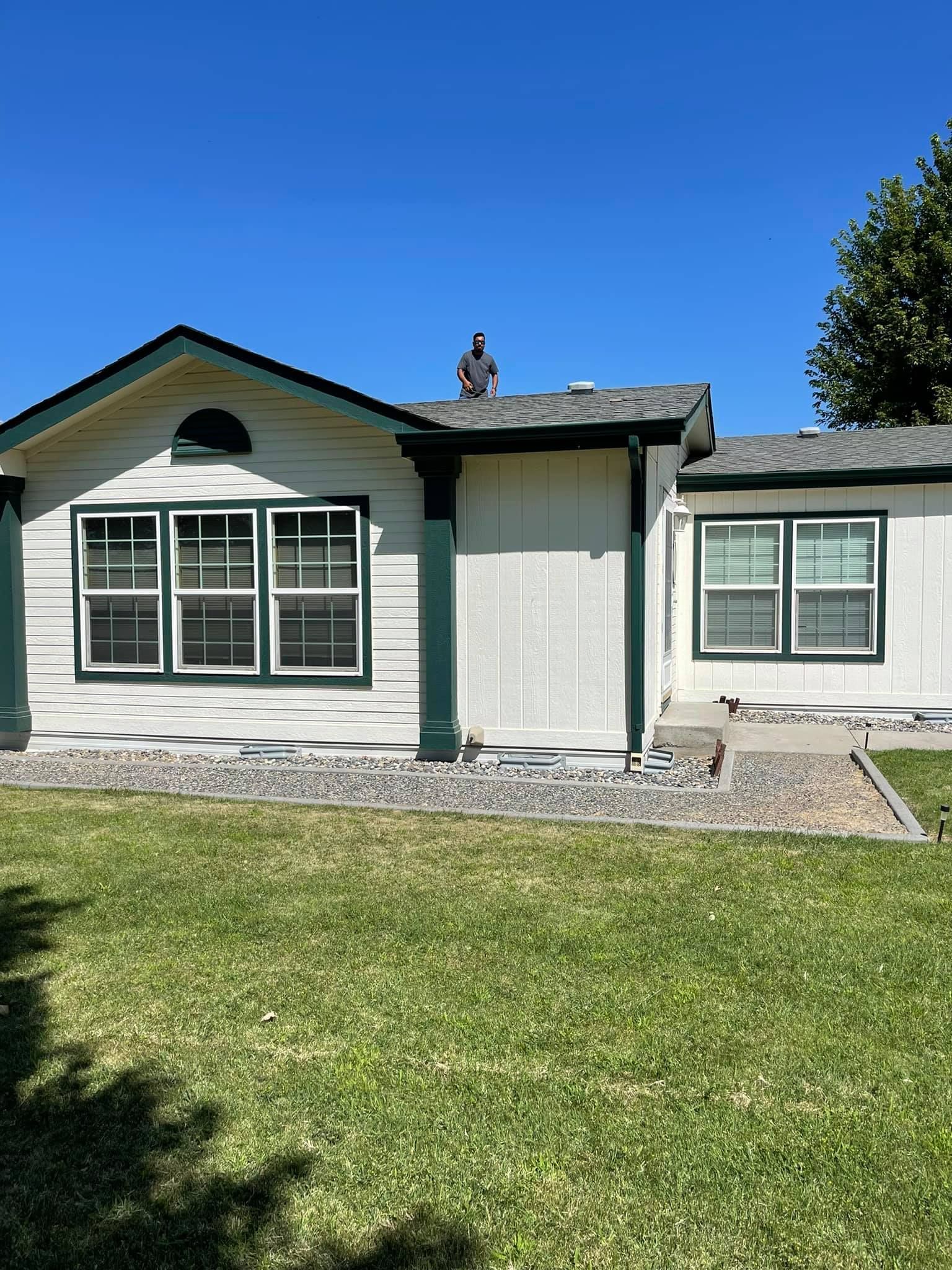 A one-story white house with green trim under a clear blue sky. A person stands on the roof.
