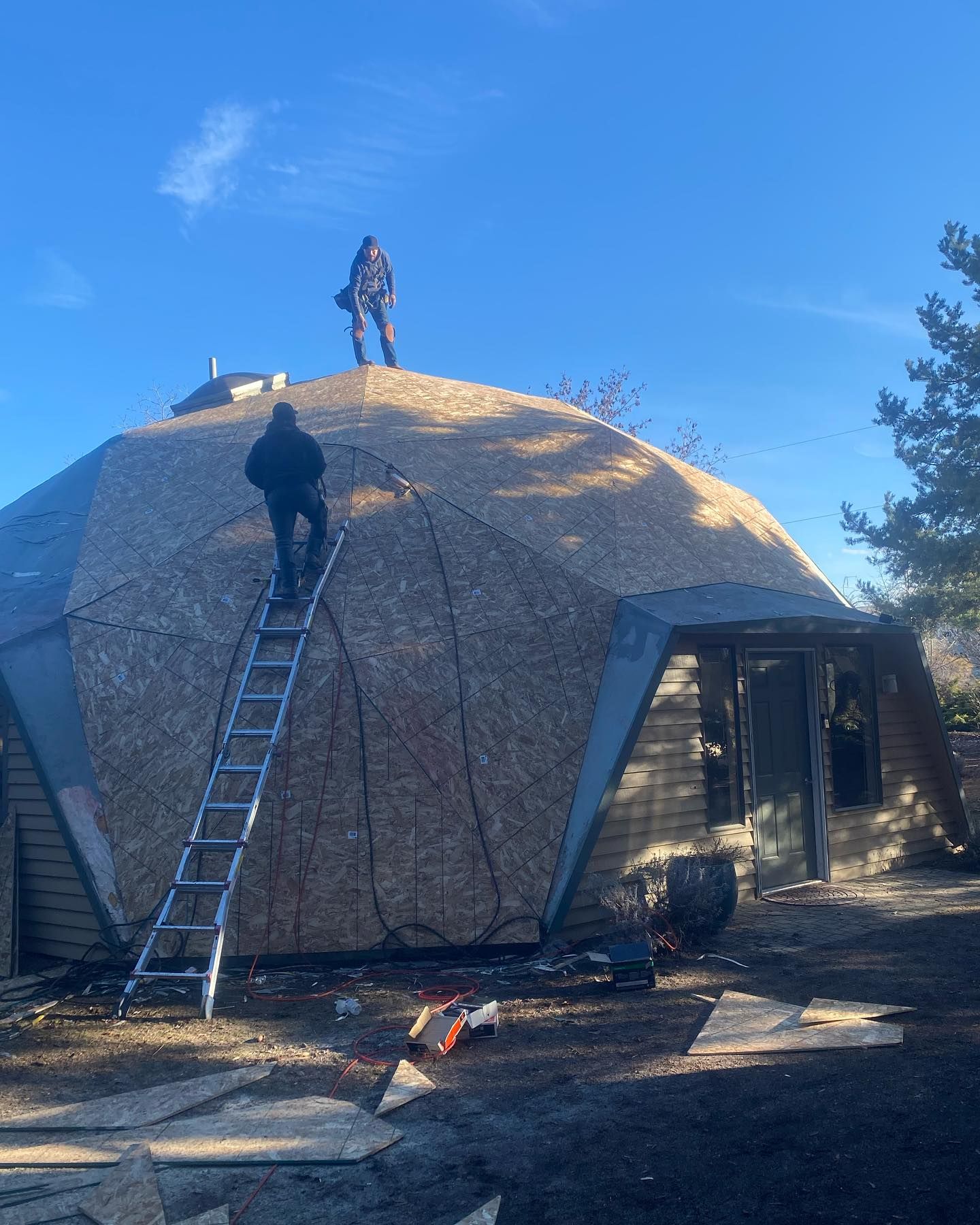 Two people on roof of geodesic dome house, one on ladder. Clear blue sky.