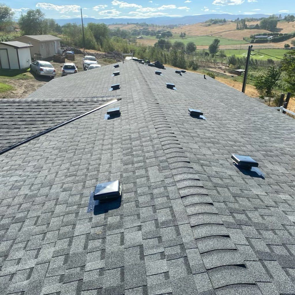 View of a dark gray shingled roof with multiple black vents, overlooking a rural landscape.