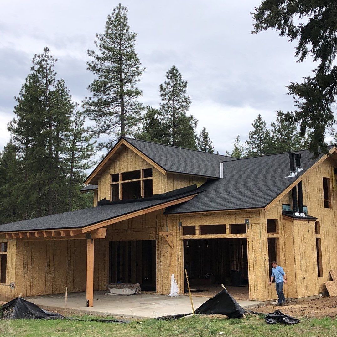 House under construction with exposed wooden frame and dark roof, set amongst trees.