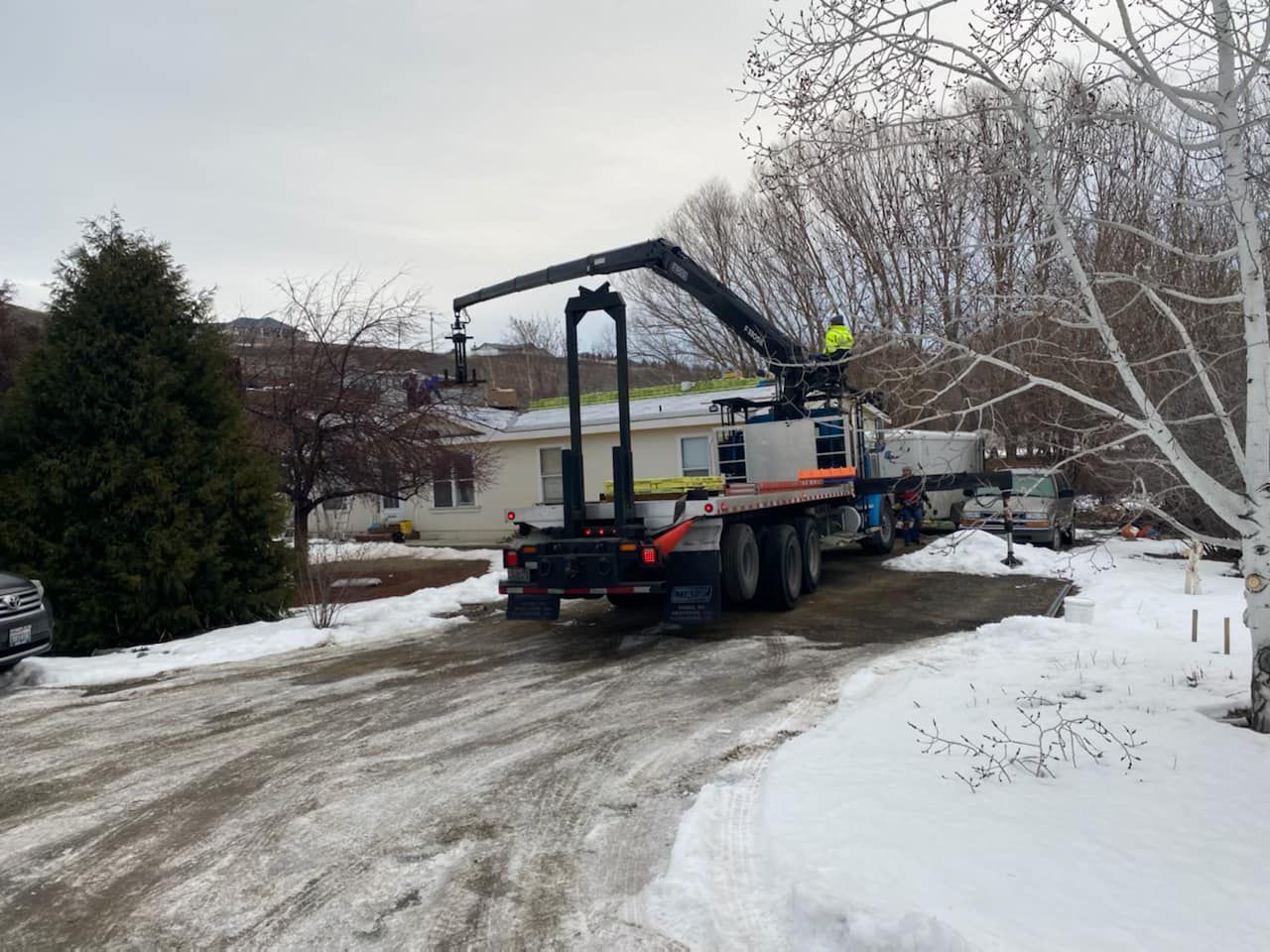 Truck with a crane lifting something onto the roof of a house; snow on the ground; worker in high-vis vest.
