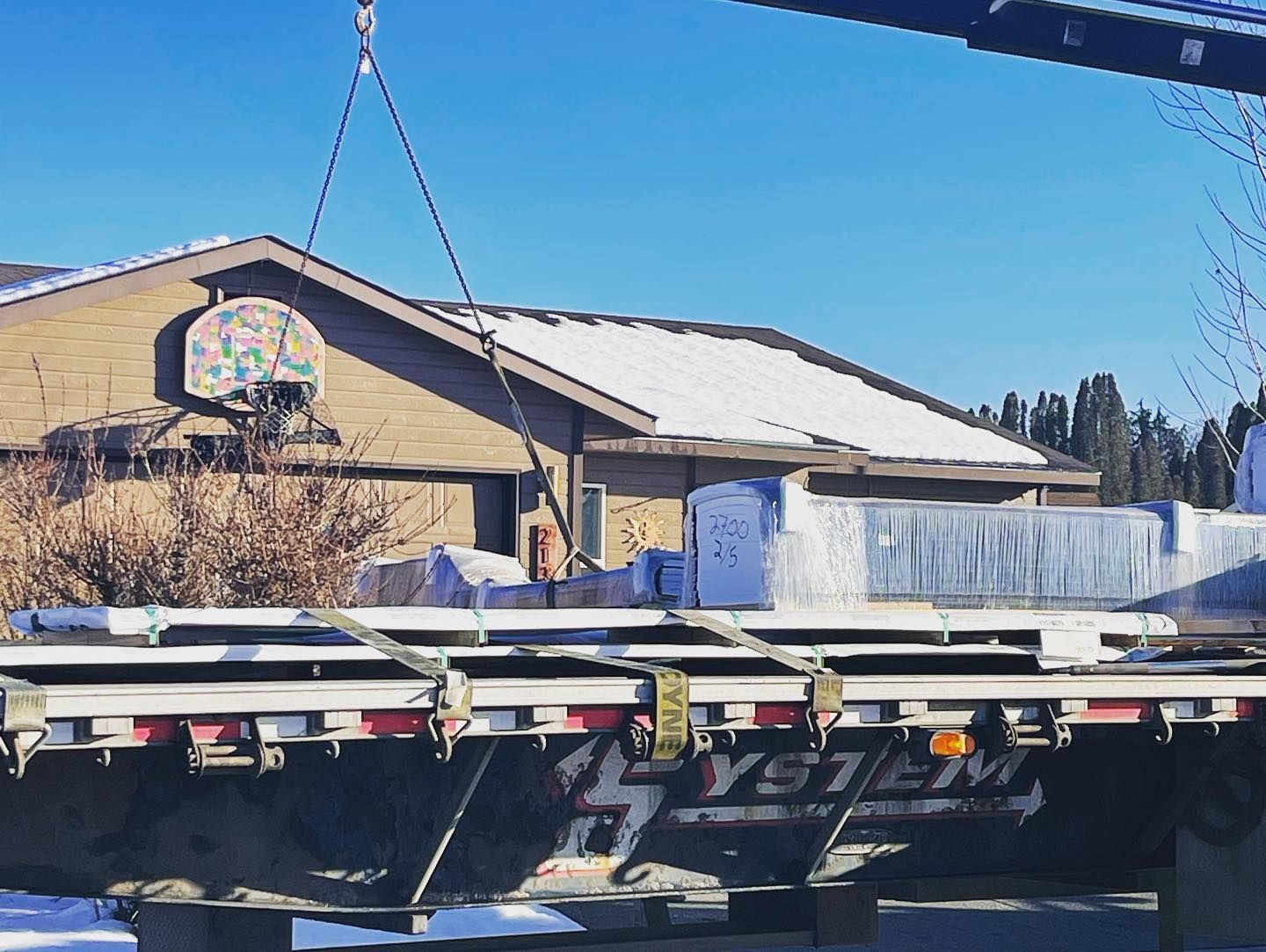 Crane lifting rectangular panels from a flatbed truck in front of a house with a snow-covered roof.