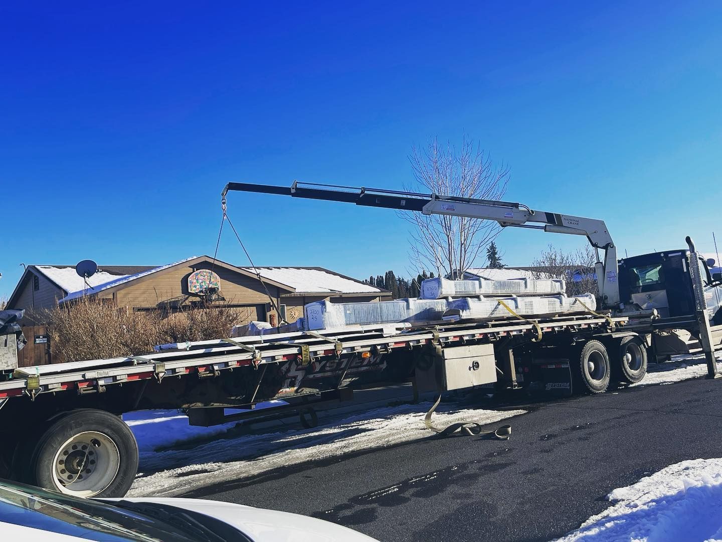 Flatbed truck with crane on a snow-covered street in front of a house, bright blue sky.