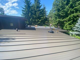 Brown metal roof of a house with trees and a blue sky in the background.