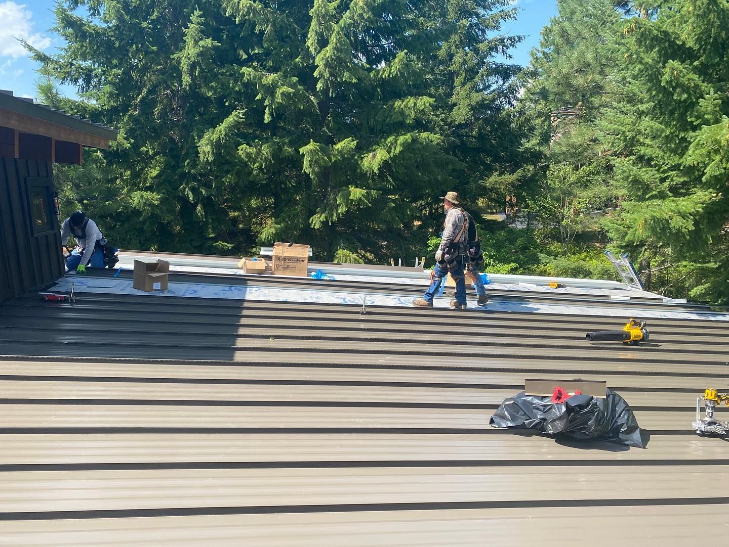 Two people on a rooftop installing material; sunny day, trees in background, tools and supplies visible.