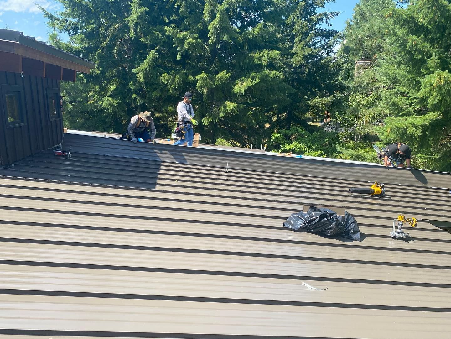 Workers installing roofing on a house, tools and trash on the roof, trees in the background.