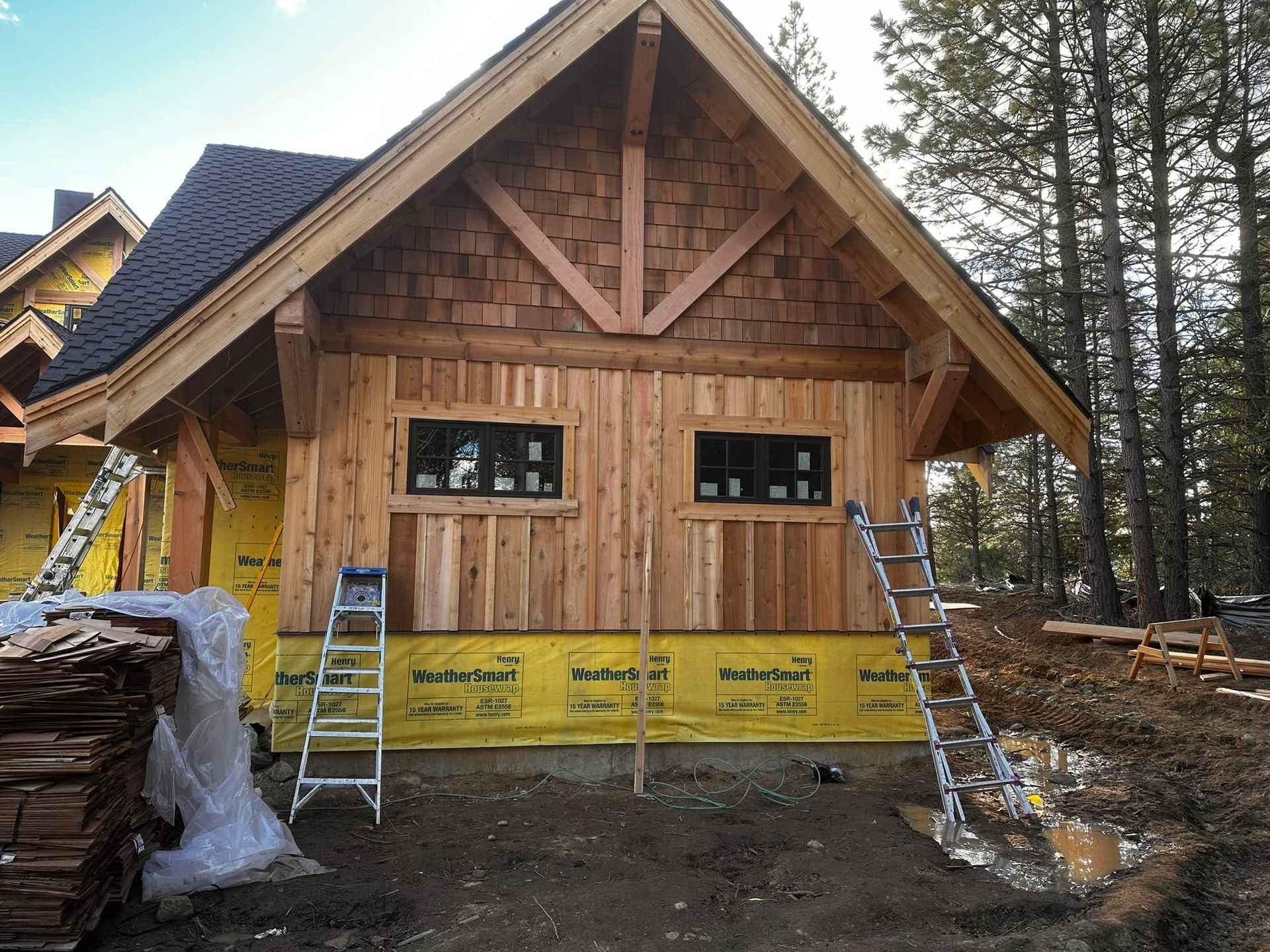 Wooden cabin under construction with two black windows, ladders, and surrounding trees.
