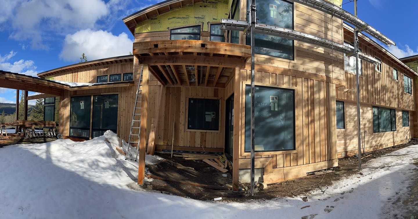 Building under construction, with wooden frame, large windows, and snow on the ground under a blue sky.