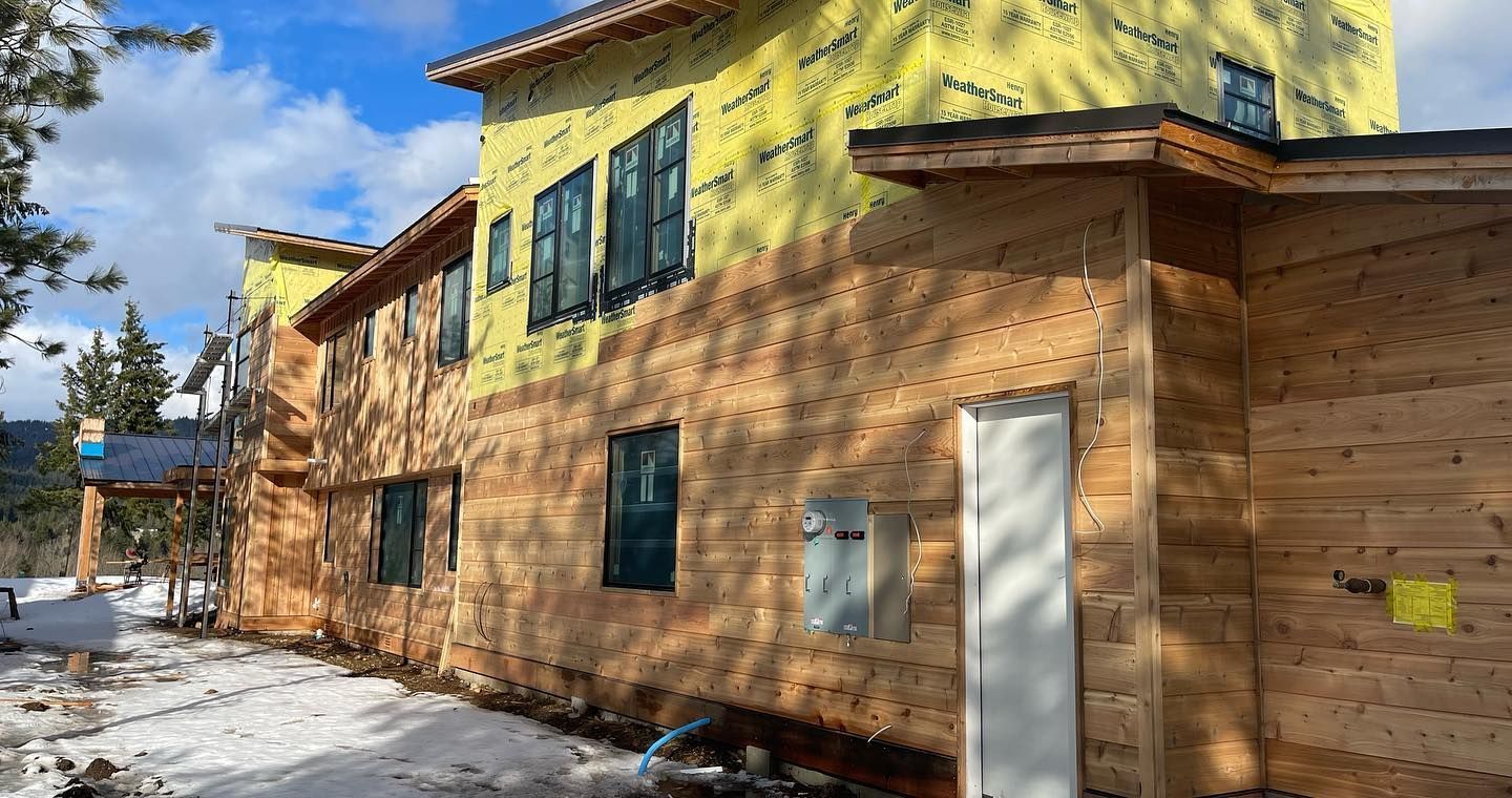 Building under construction, exterior view, with wooden siding and yellow sheathing. Snowy ground and blue sky.
