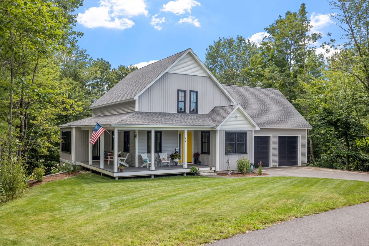 Gray house with porch, American flag, yellow door, green lawn, trees, blue sky.