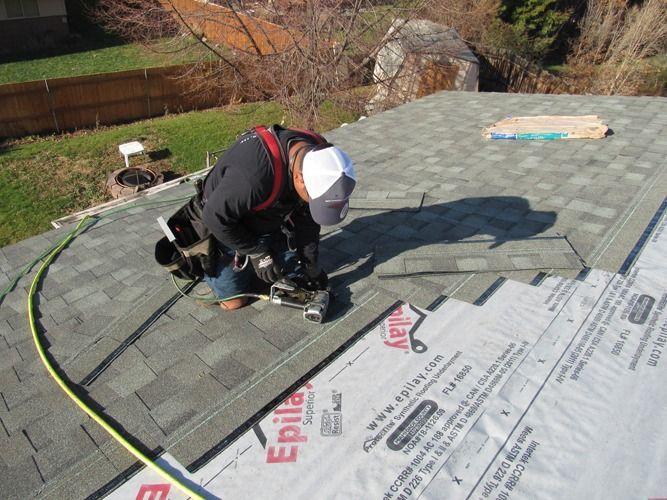 Roofer on a residential roof, installing shingles. Gray and white roofing materials, green hose, sunny day.