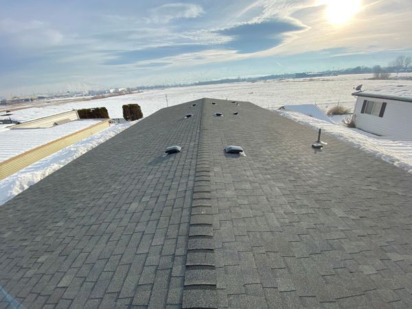 View of a gray shingled roof with three vents, surrounded by snow-covered ground under a partly sunny sky.