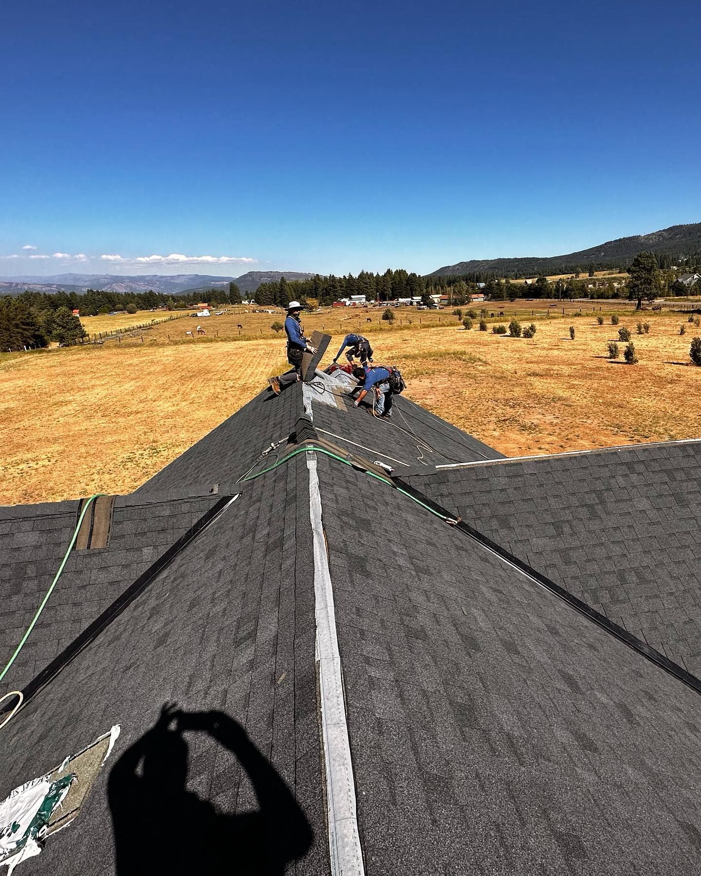 Roofers working on a black asphalt shingle roof under a blue sky, overlooking a field.