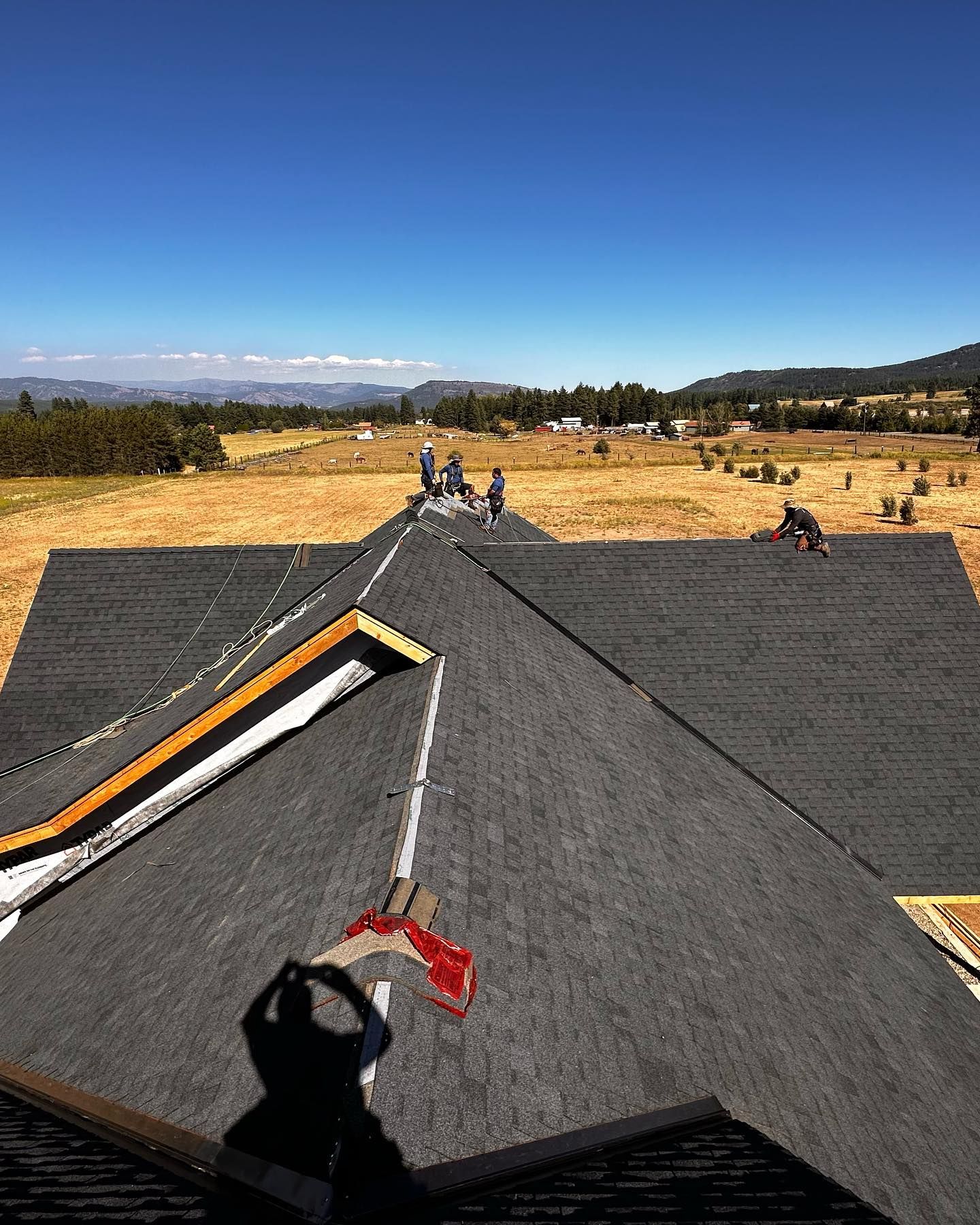 Roofers installing shingles on a house roof under a blue sky, with a field and mountains in the background.
