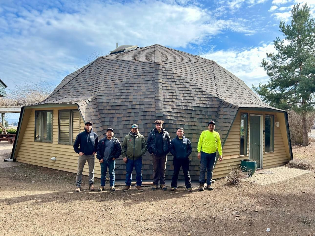 Group of men in front of a dome-shaped building. Cloudy sky, brown shingles, beige siding.