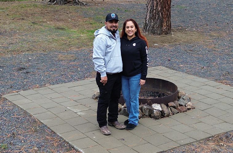 Couple standing near a fire pit on a stone patio; man in hoodie and jeans, woman in black sweatshirt and jeans.