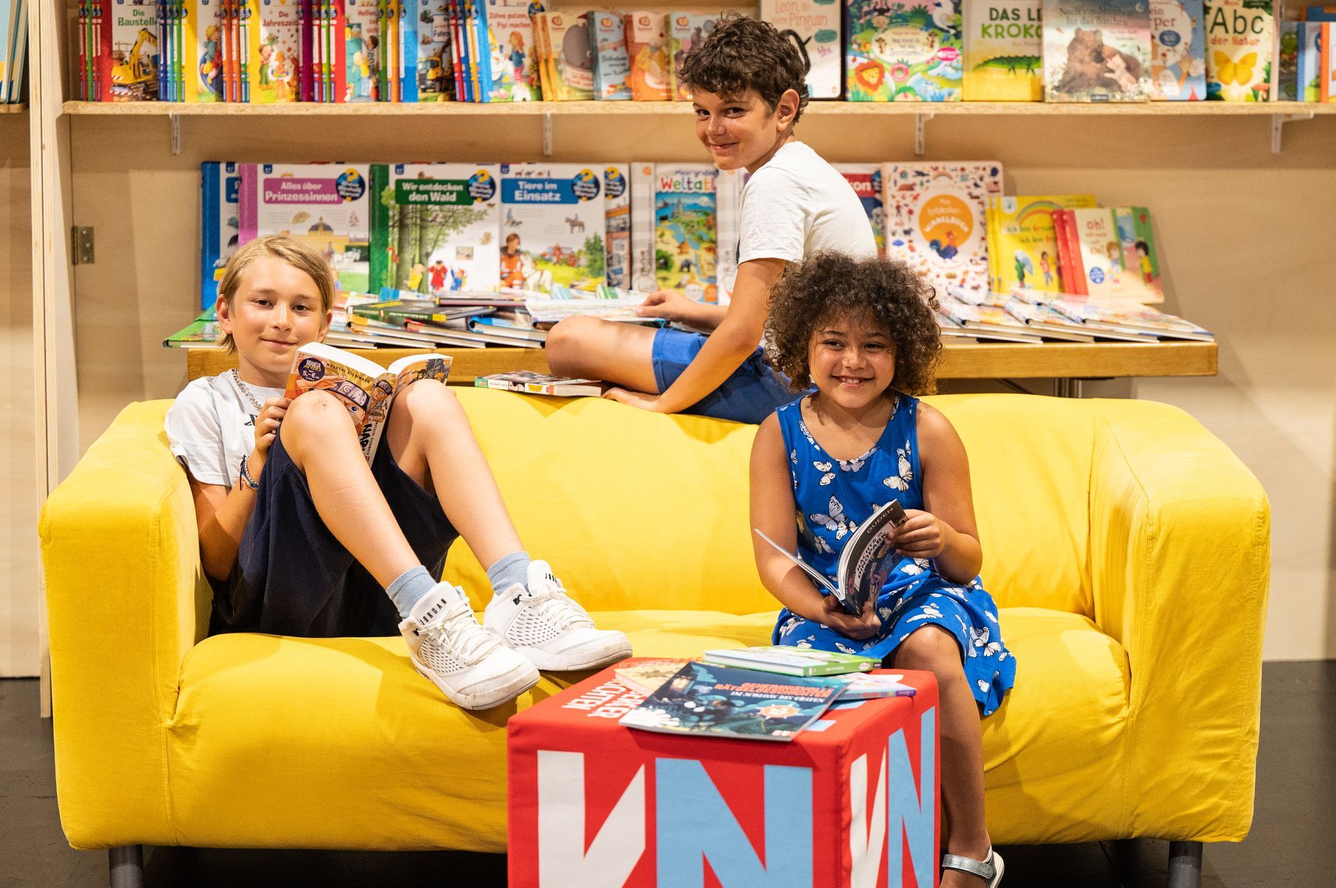 Drei Kinder sitzen auf einem gelben Sofa in einer Bibliothek.