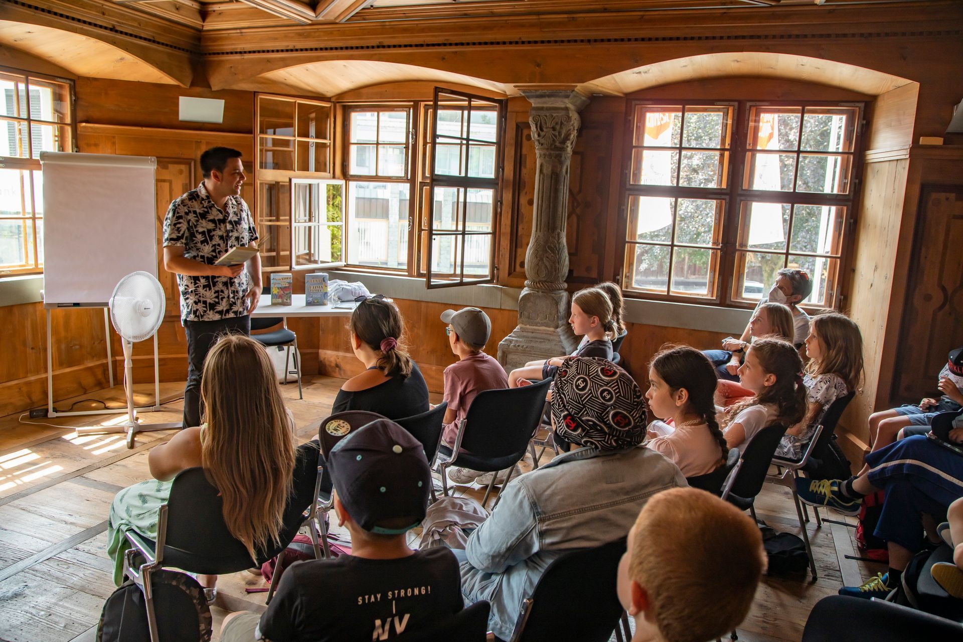 Eine Gruppe von Menschen sitzt in einem Raum auf Stühlen, vor ihnen steht ein Mann.