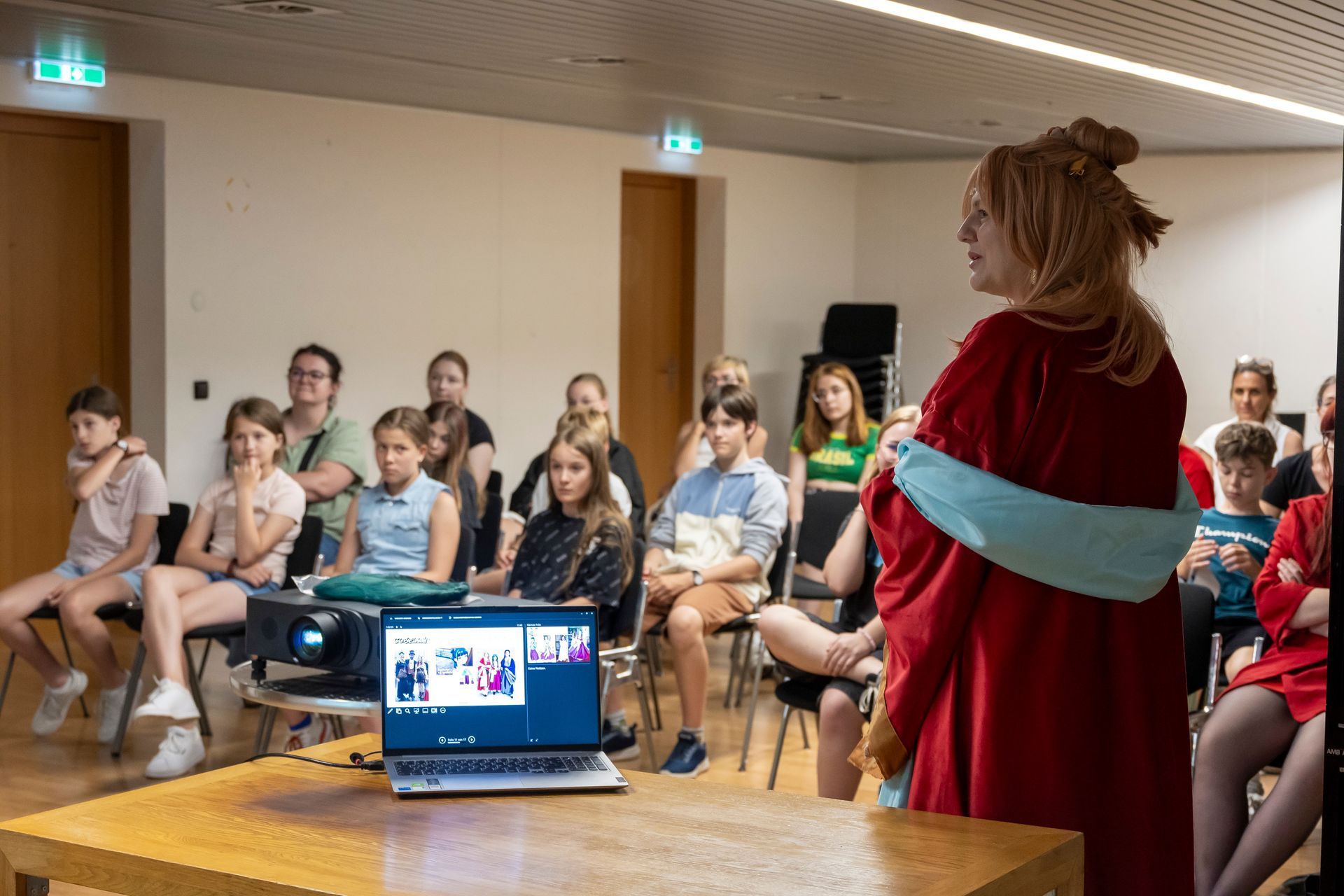 Eine Frau in einem roten Gewand steht vor einer Gruppe von Menschen, die auf Stühlen sitzen.
