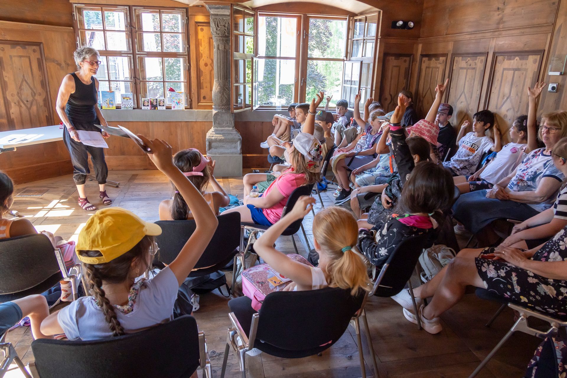 Eine Gruppe von Menschen sitzt mit erhobenen Händen auf Stühlen in einem Raum.