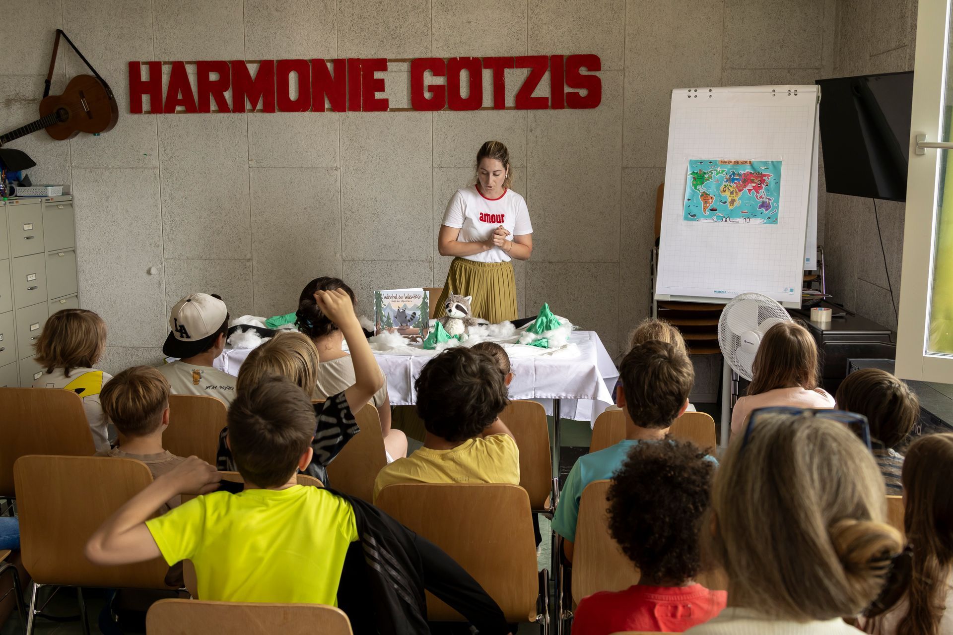 Eine Frau hält vor einem Schild mit der Aufschrift „Harmonie Götzis“ einen Vortrag vor einer Gruppe von Kindern.