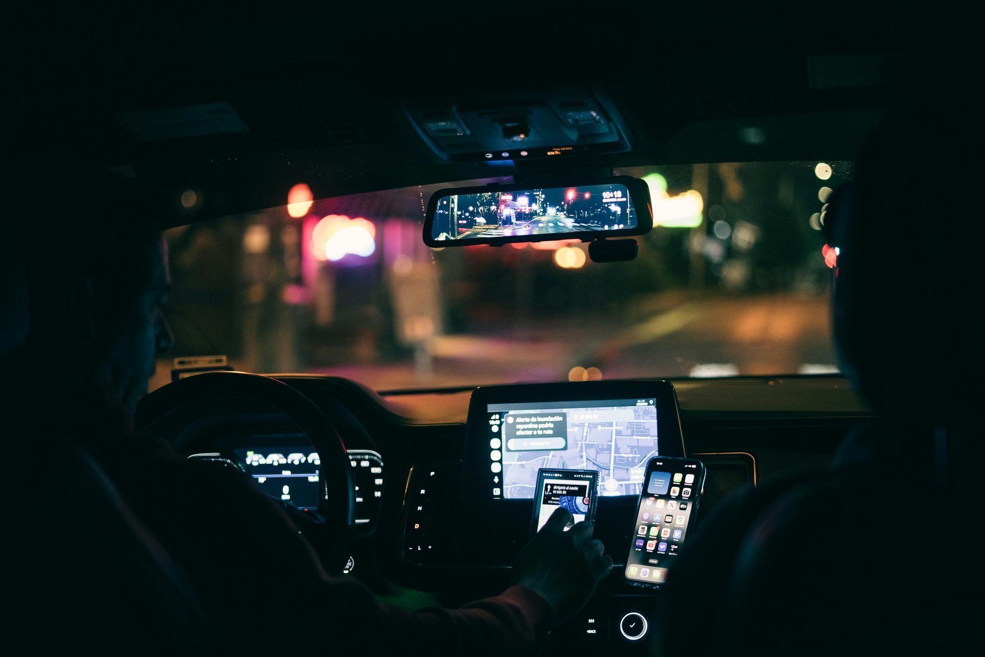 Inside a car at night, two people use multiple phones while navigating. City lights visible outside.
