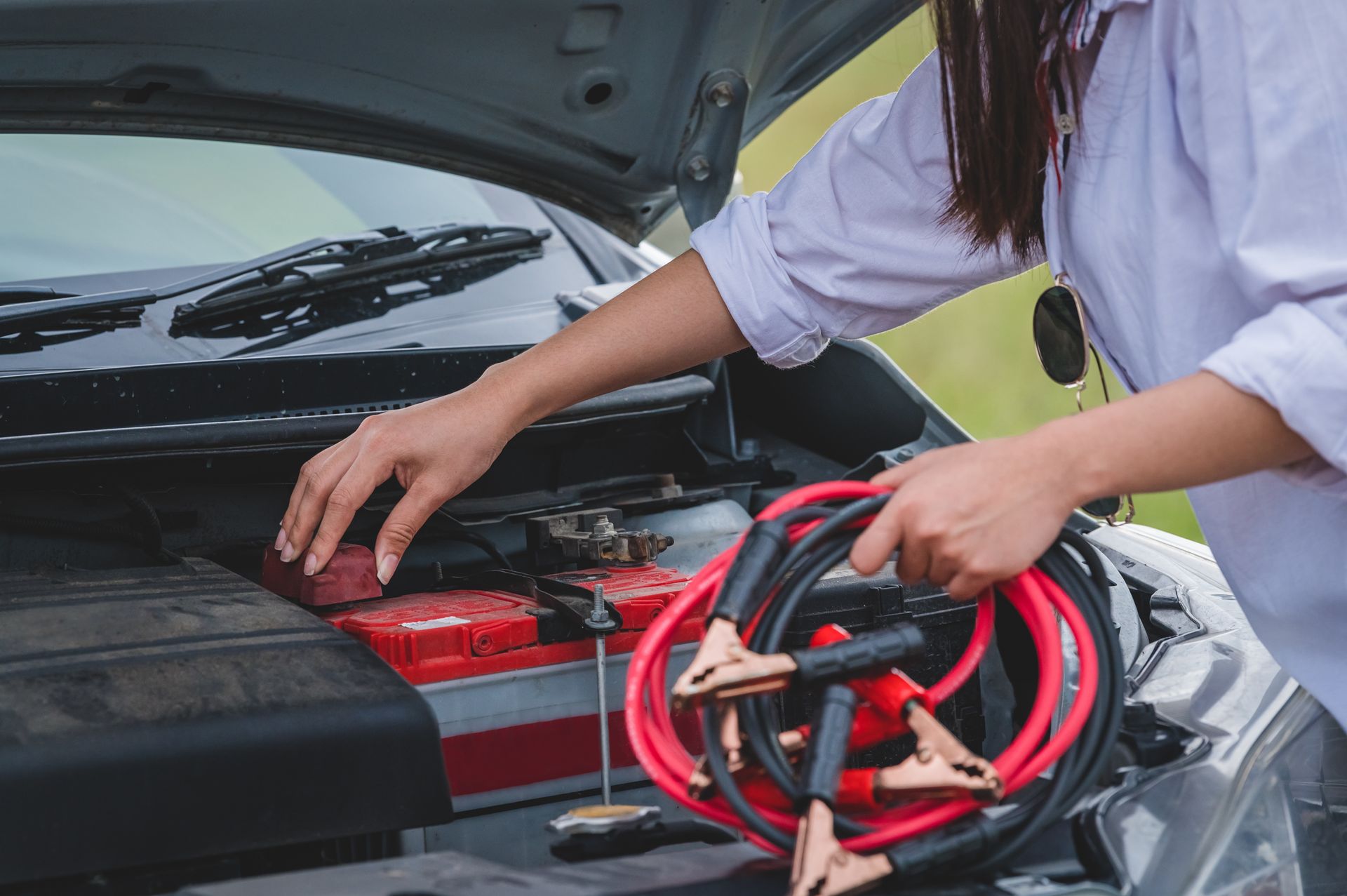 A person holding jumper cables prepares to attach them to a car battery under the hood of an open vehicle.