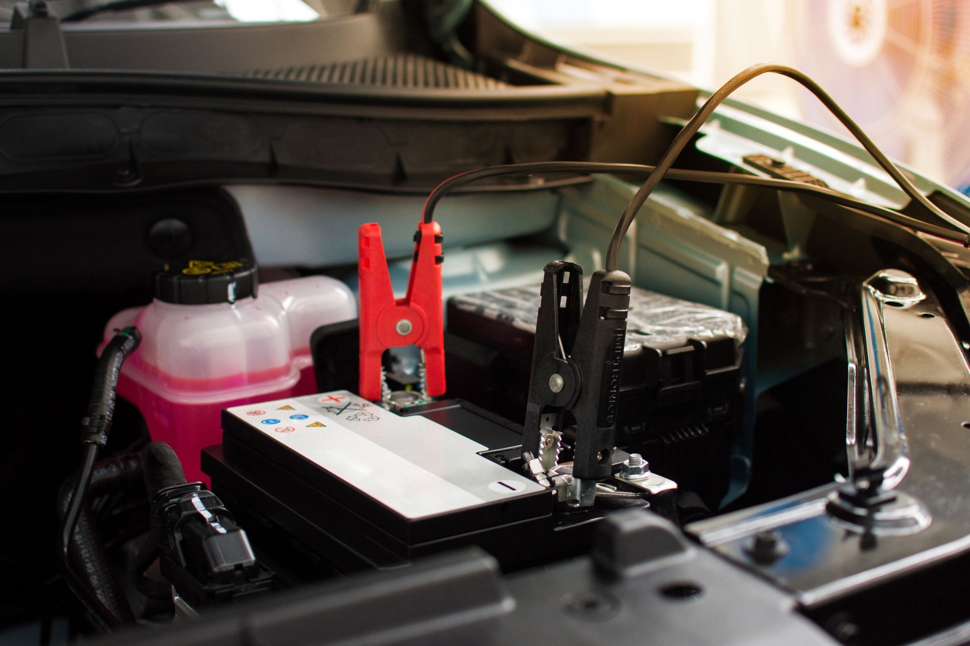 A car battery in an engine bay with red and black jumper cable clamps attached to the terminals.