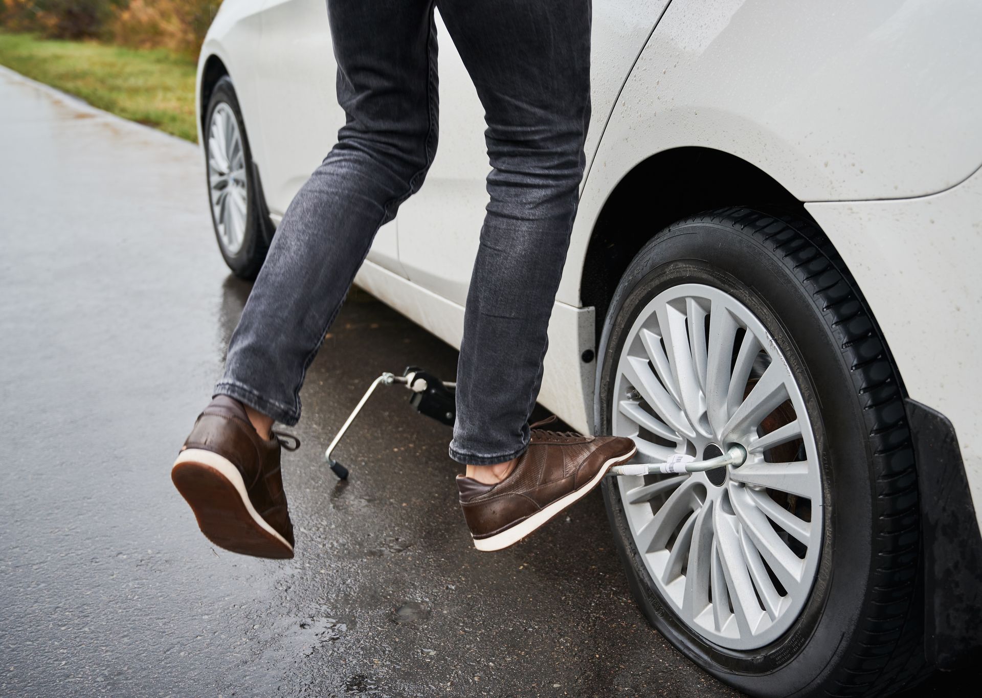 A person standing on a wet road uses their foot to press a tire iron against a lug nut on a car with a flat tire.
