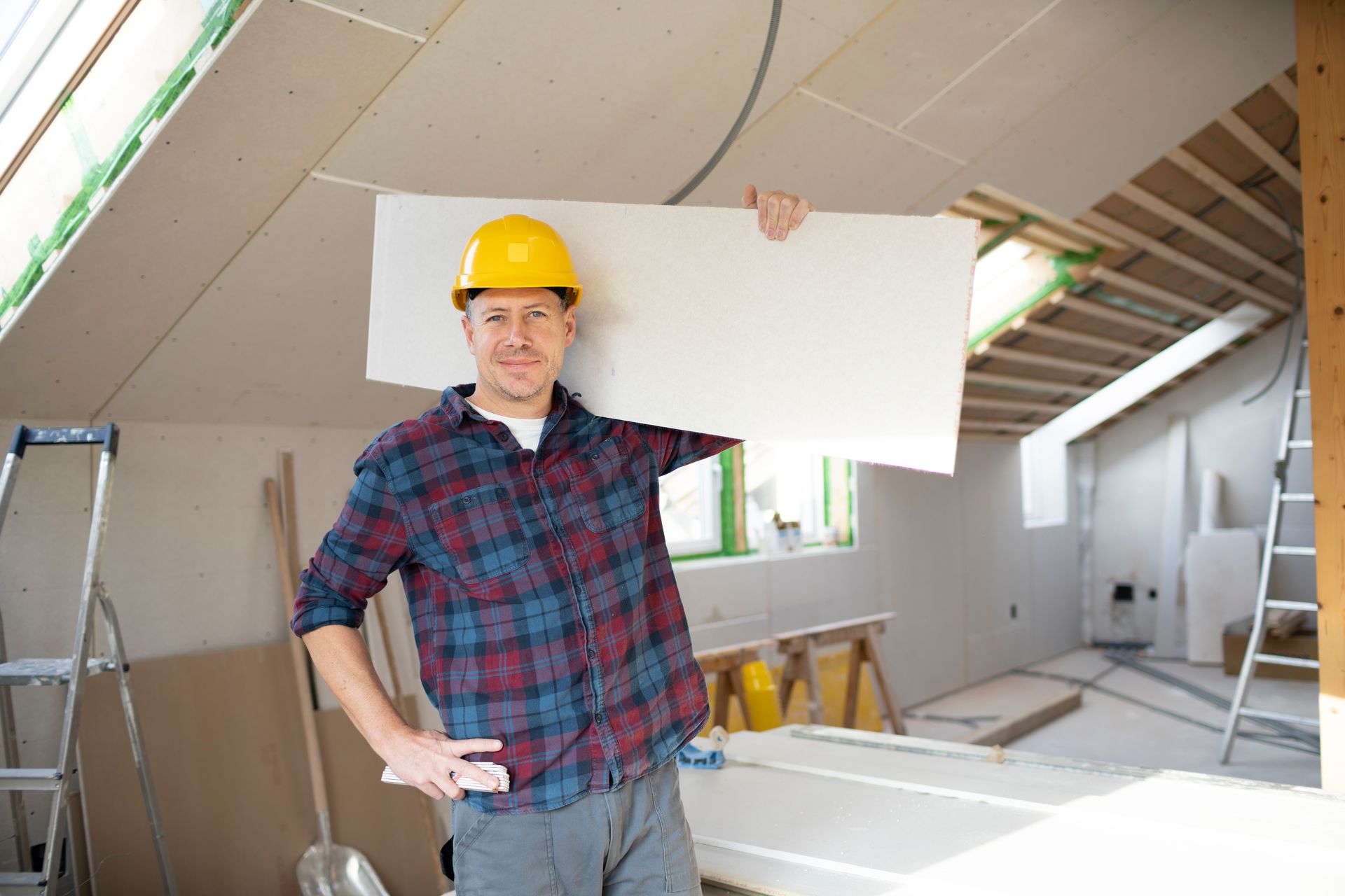 Construction worker in yellow hard hat holding drywall in unfinished room.