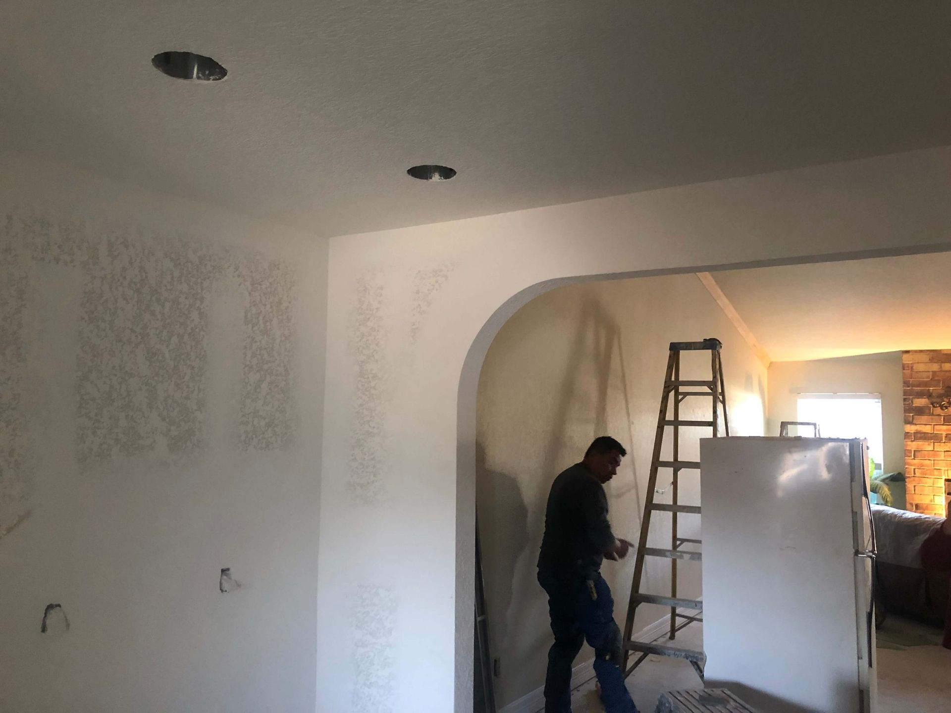 Man installing a light fixture in a ceiling. White walls, archway, and ladder visible.