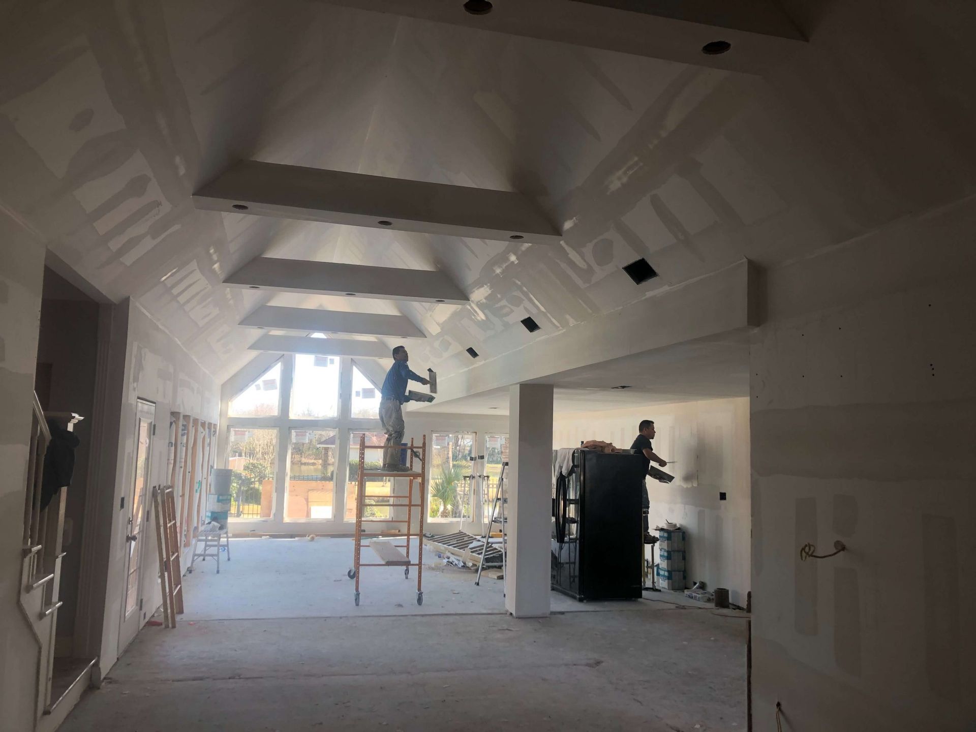 Workers drywalling a room with a vaulted ceiling and skylights. Construction materials are scattered about.