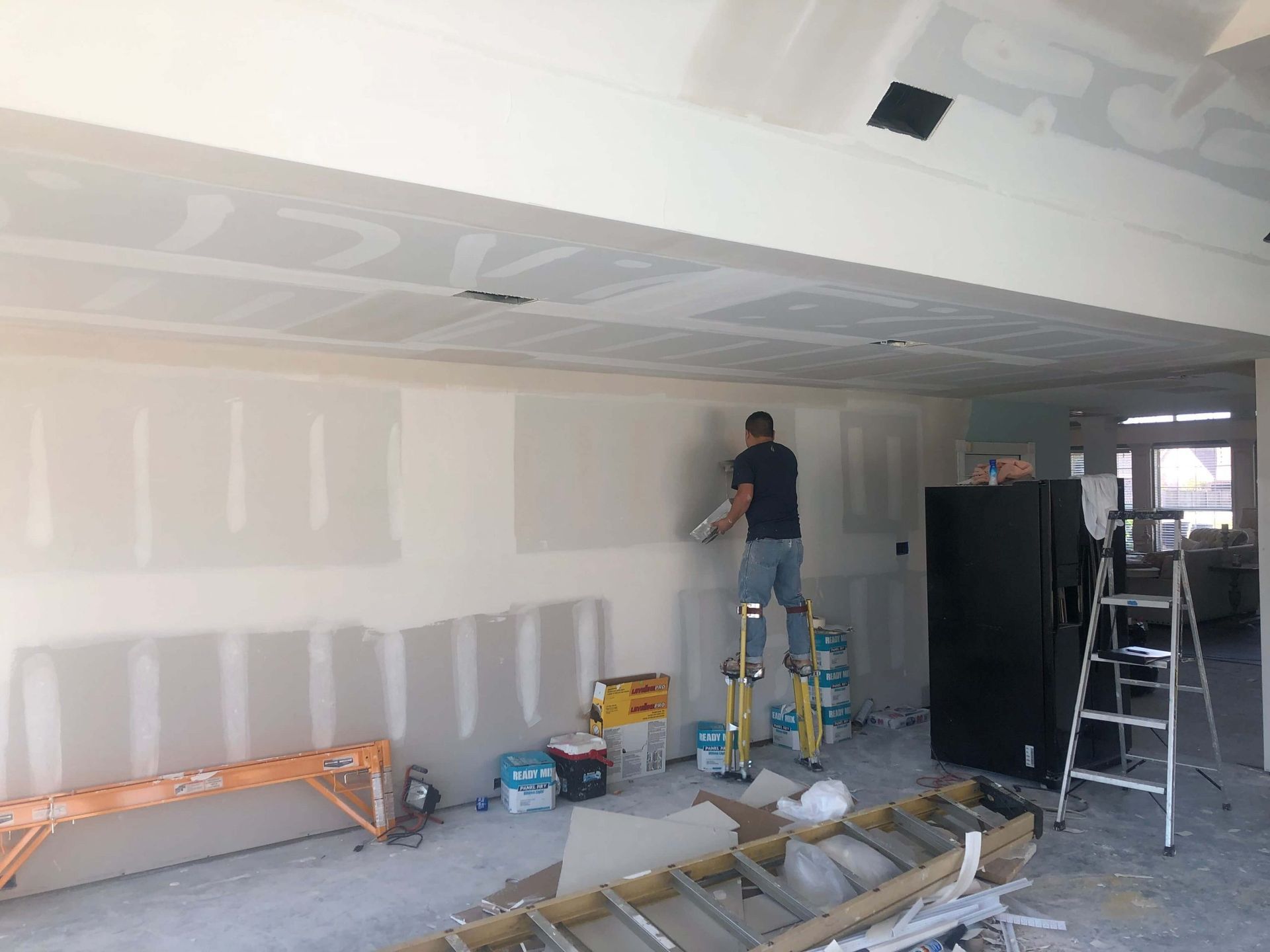 Man on a ladder applying drywall compound to a wall in a room under construction.
