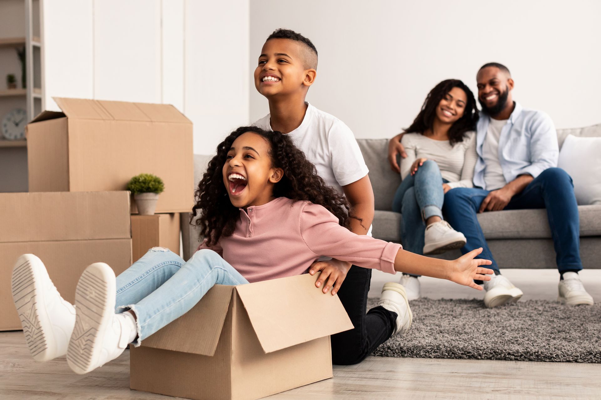 Family in a new home, girl smiles while riding in a box, siblings laugh, parents watch, boxes on the floor.
