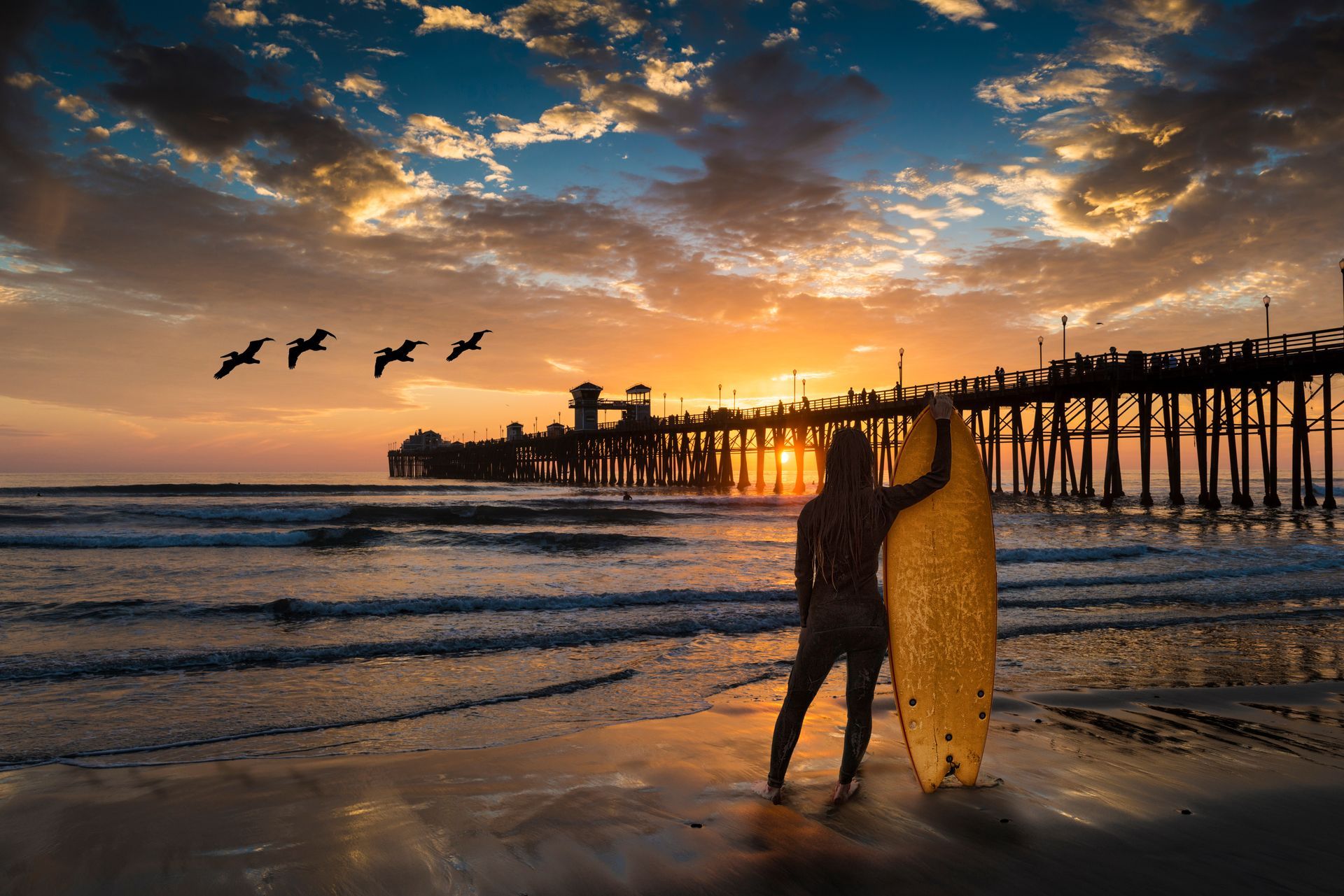 Surfer on beach watching sunset; pier silhouette, pelicans flying. Yellow surfboard, ocean waves, orange sky.
