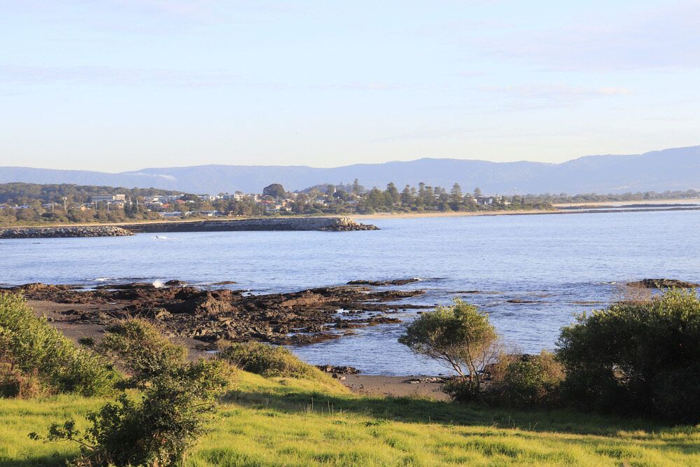 Coastal view with calm water, rocky shore, green grass, town in the distance, and mountains under a blue sky.